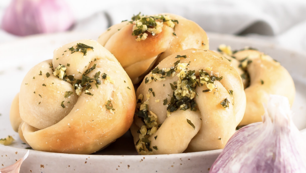 Sourdough starter discard garlic knots are neatly stacked on a white plate, garnished with a parsley and garlic oil concoction. Warm and ready to be enjoyed.