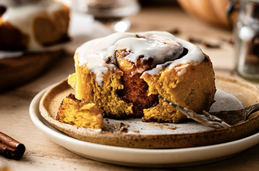 Sourdough starter discard pumpkin roll sitting on a plate with a fork with one bite missing; topped with a white drizzle that is melted and running down the sides.