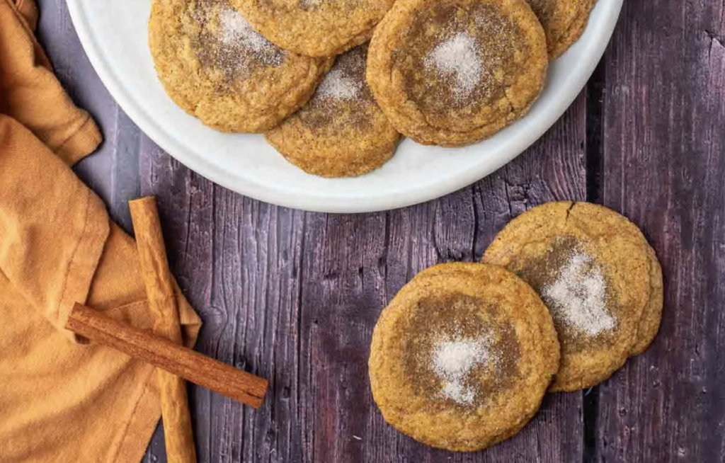 Sourdough starter discard pumpkin maple cookies sitting on a plate and table with cinnamon sticks, topped with sugar in the middle; warm, gooey and ready to be enjoyed.