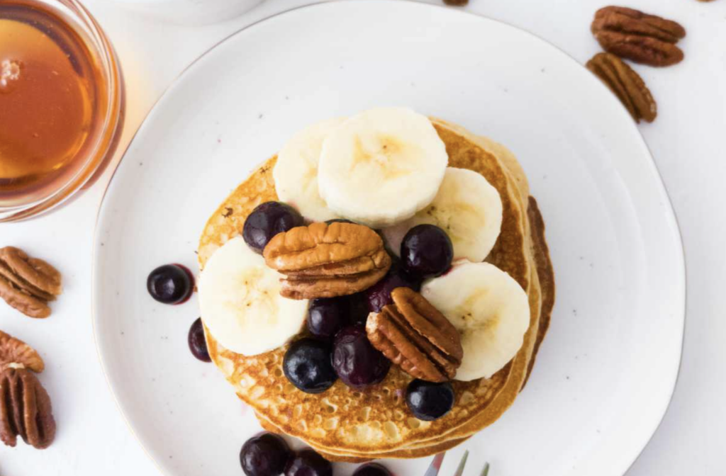 Sourdough starter discard banana pancakes sitting on a white pate, topped with pecans, bananas, blueberries and syrup.