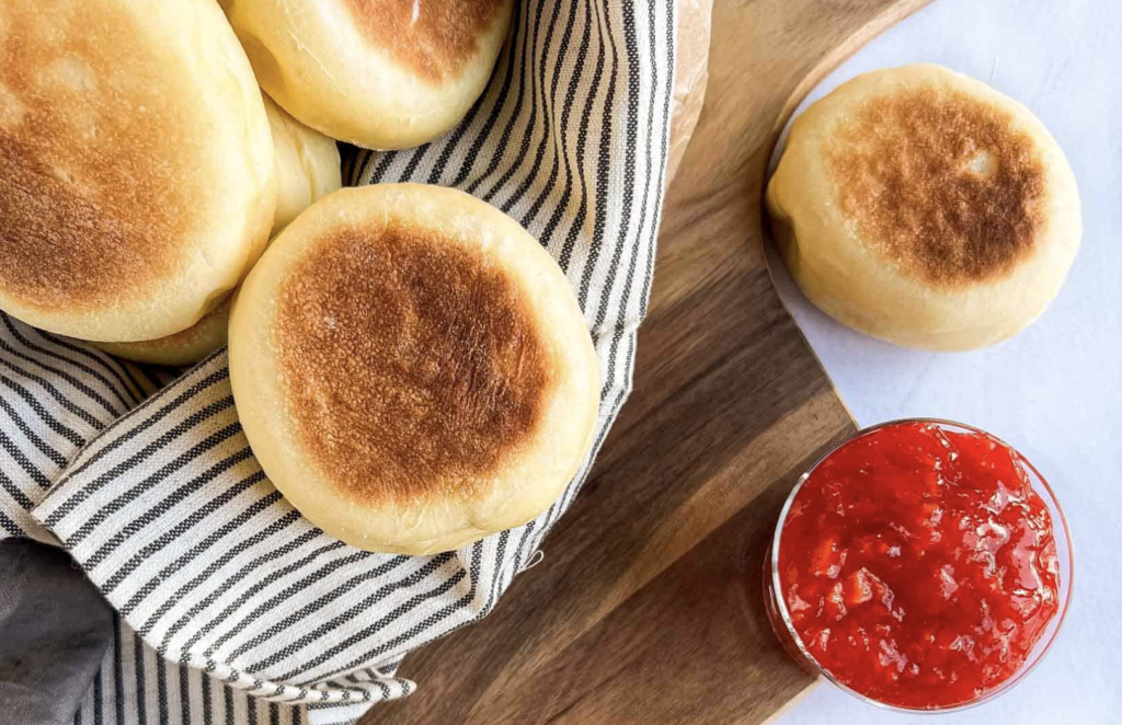 Sourdough starter discard english muffins aesthetically staged on a wooden cutting board with a red jam of sort on a small dish next to them, ready to be enjoyed.