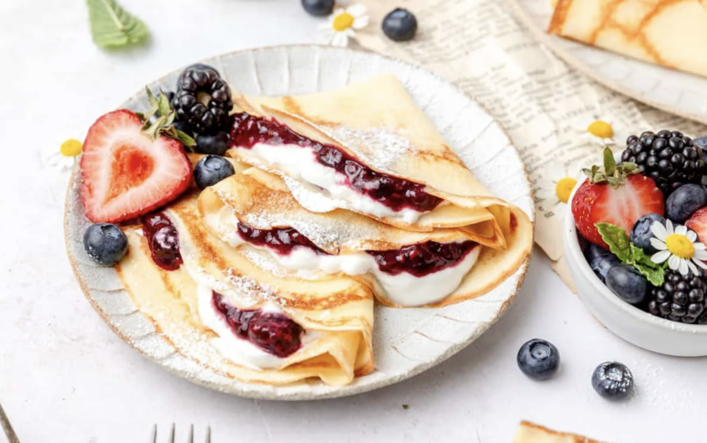 Sourdough starter discard crepes sitting on a white plate with strawberries, blueberries, blackberries, whipped cream and powdered sugar, ready to be enjoyed.