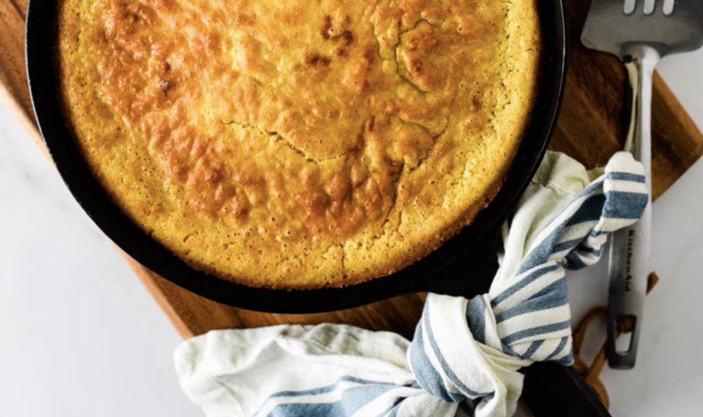 Sourdough starter discard cornbread cooling and sitting in a round cast iron skillet, aesthetically stages on a wooden cutting board with a blue and cream towel.