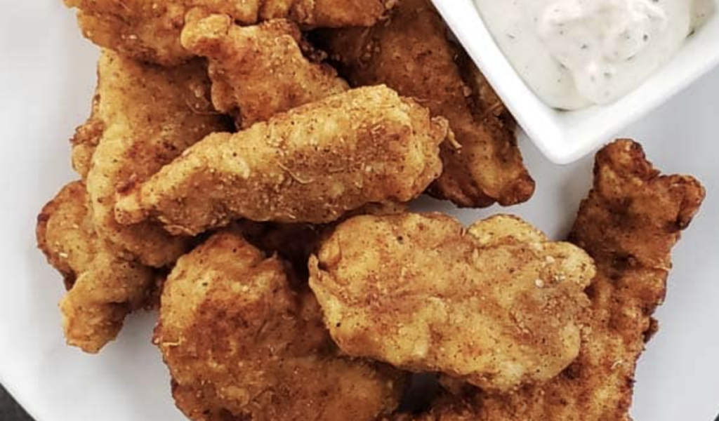 Sourdough starter discard battered and fried chicken sitting in a stack on a white plate next to a small, white bowl of homemade ranch.