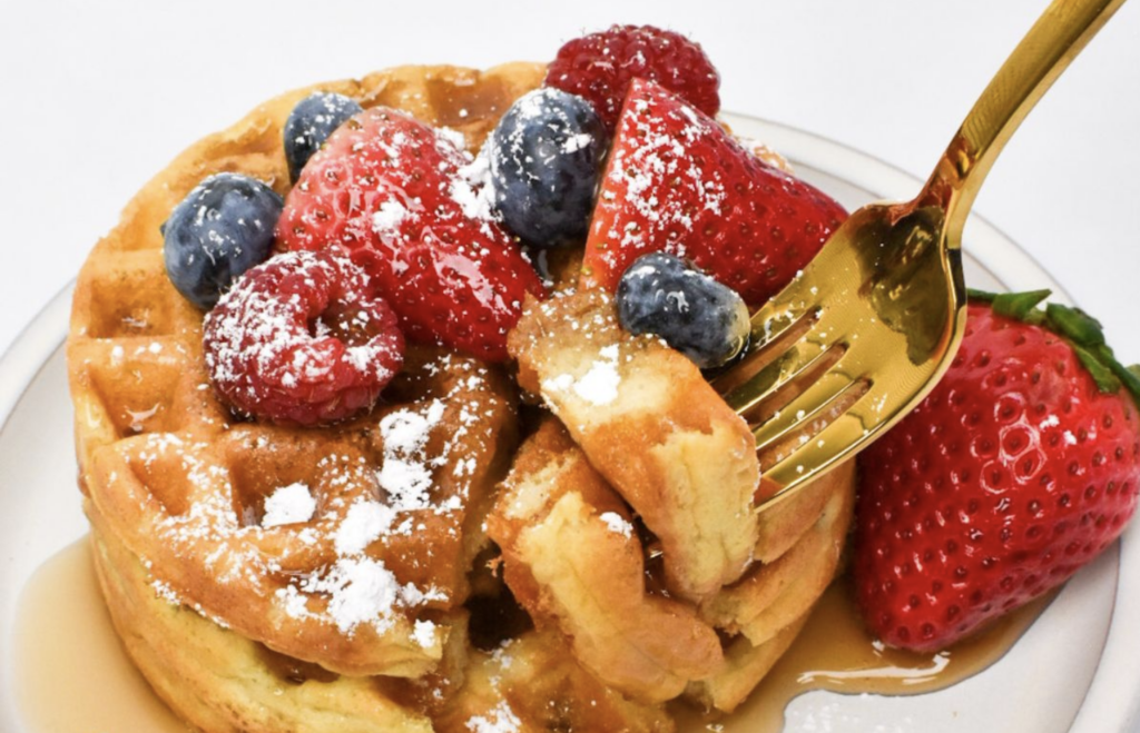 Two sourdough starter discard waffles stacked on a white plate, topped with blueberries, strawberries, raspberries and powdered sugar; a golden fork is approaching the stack ready to take a bite.