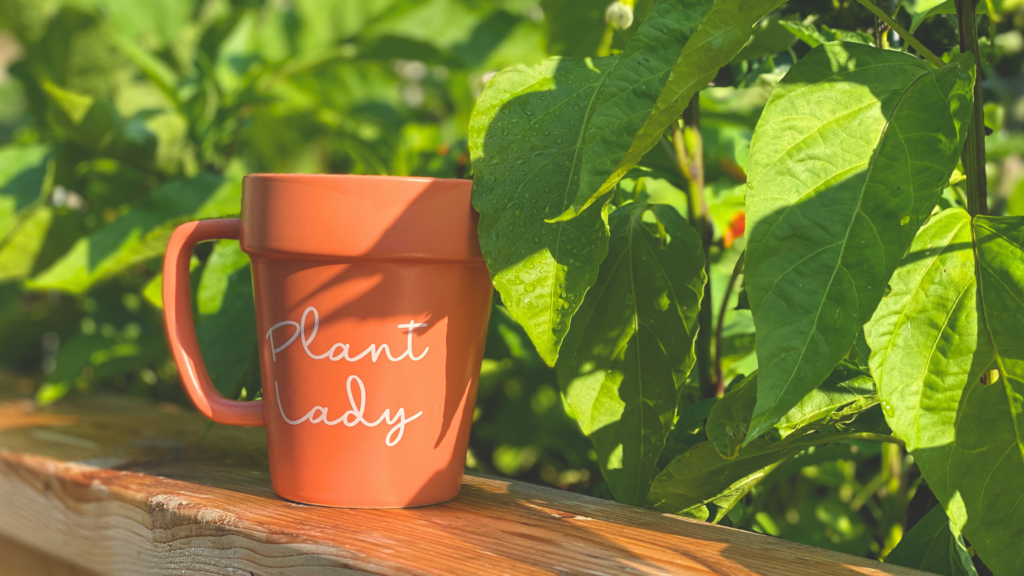 orange coffee mug with "plant lady" written on the front staged on the top of a garden bed under green foliage