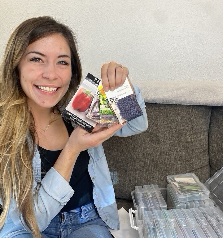 Woman holding vegetable seed packets in both hands posing for a staged photo with seed container sitting on couch next to her