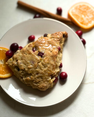 image of cranberry orange scone on a white plate surrounded by cranberries and cinnamon sticks